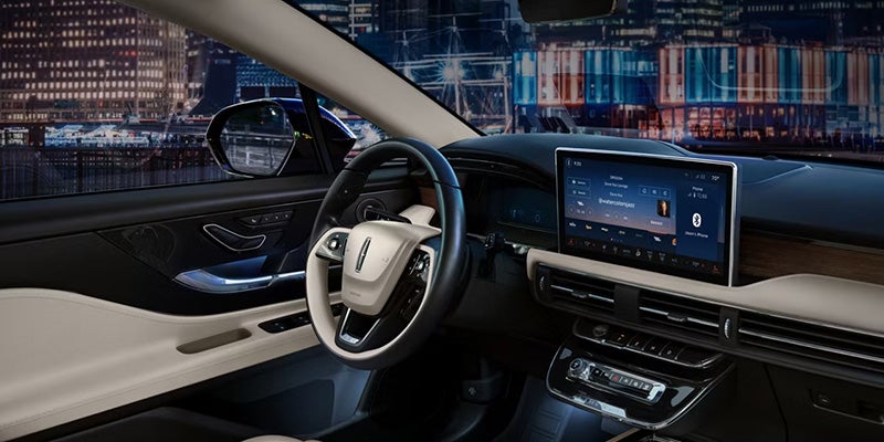 Modern car interior at night with a steering wheel, large infotainment screen, and city lights visible outside.