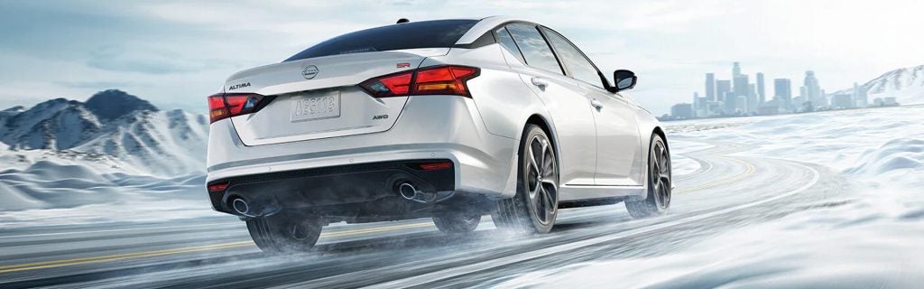 Rear three-quarter view of a white Nissan Altima SR AWD driving on a snow-covered road with snow spraying from the tires.