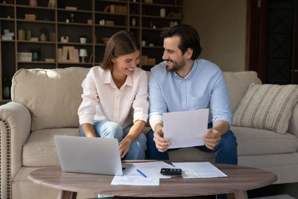 A couple sitting on a couch, reviewing paperwork together with a laptop and calculator on the table.