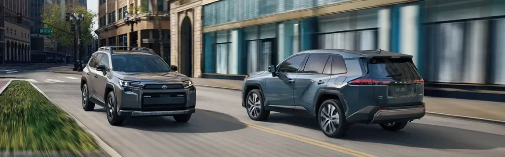 Two Toyota SUVs, a gray Toyota RAV4 and a silver Toyota RAV4, driving side by side through an urban street.