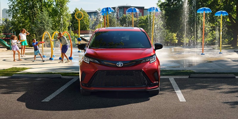 2025 Toyota Sienna in a parking lot with a family enjoying in a playground, featuring water fountains and vibrant blue umbrellas in the background.