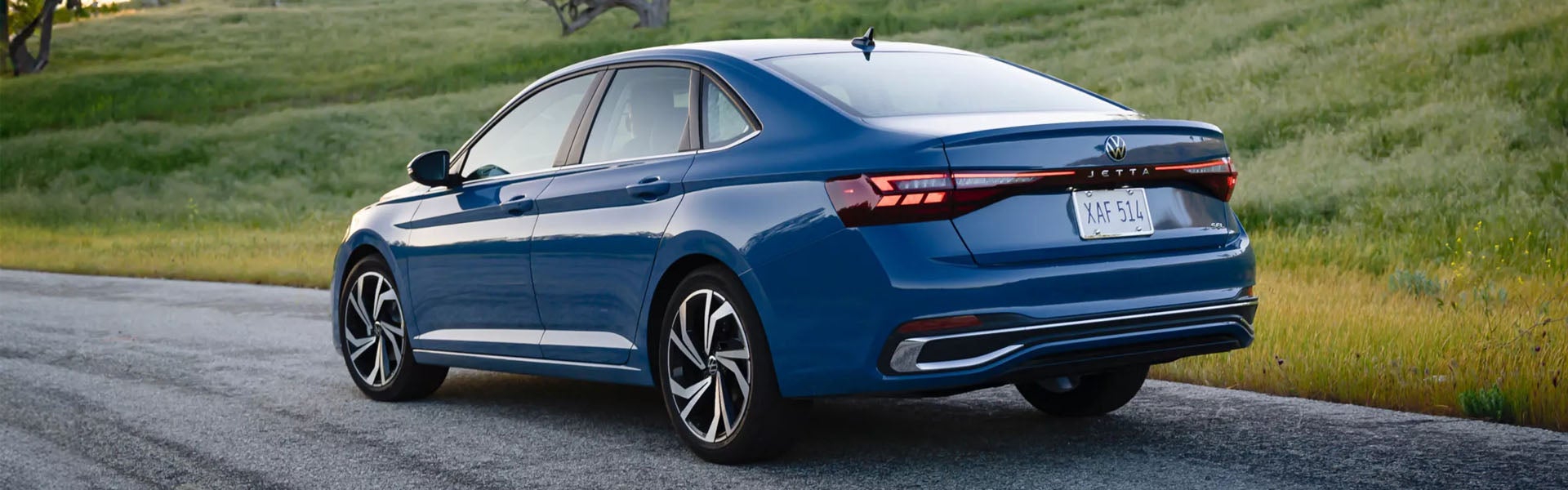 2026 Volkswagen Jetta driving on a scenic road, showcasing rear design, LED taillights, and sleek blue sedan styling.