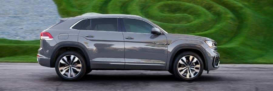 a Volkswagen Atlas Cross Sport in pure grey drives down a paved road with a landscaping feature behind it