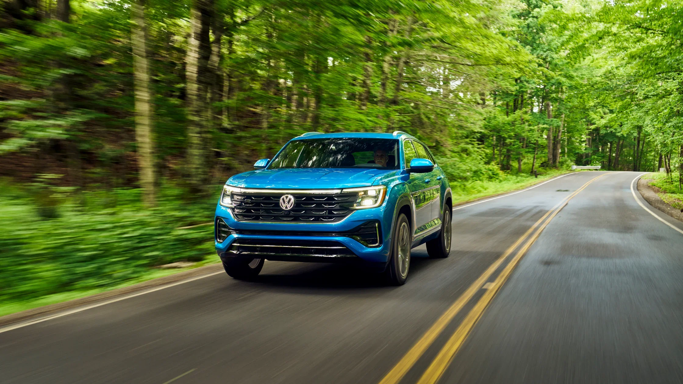 2024 Volkswagen Atlas Cross Sport in vibrant blue driving through lush greenery.