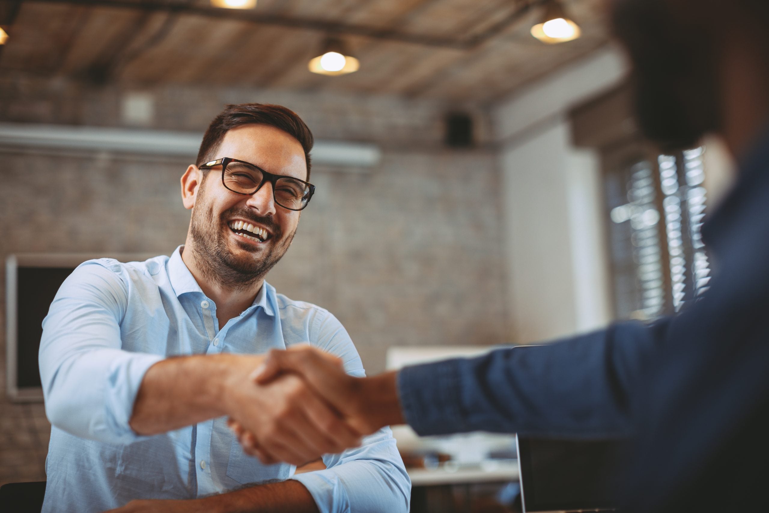 A professional smiling man shaking hands with another person in a business setting.
