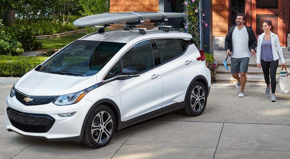 A couple is walking toward a white 2021 Chevy Bolt EV parked in a driveway with a paddle board on top.
