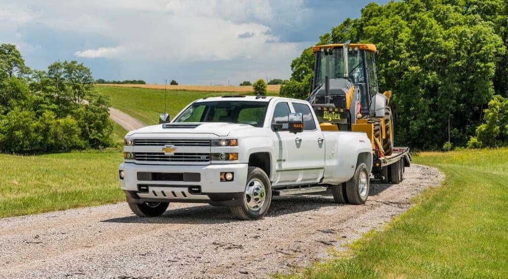 A white 2018 Chevy Silverado 3500HD is towing construction equipment on a dirt road.