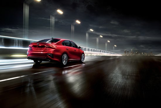 Kia car drives swiftly across a wet, illuminated bridge at night, with city lights visible in the distant background.