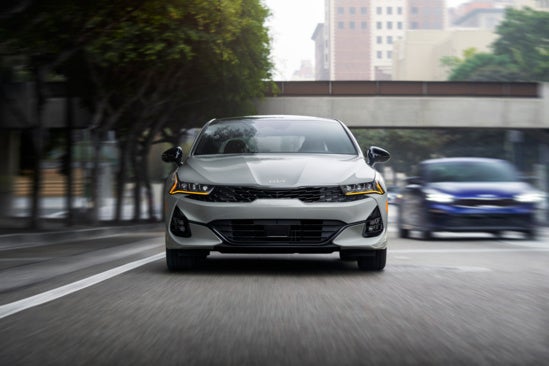 Kia K5 is driving down a city street, with blurred motion suggesting speed.