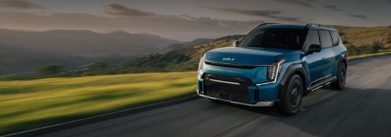 2026 KIA EV9 drives along a scenic road with rolling hills.