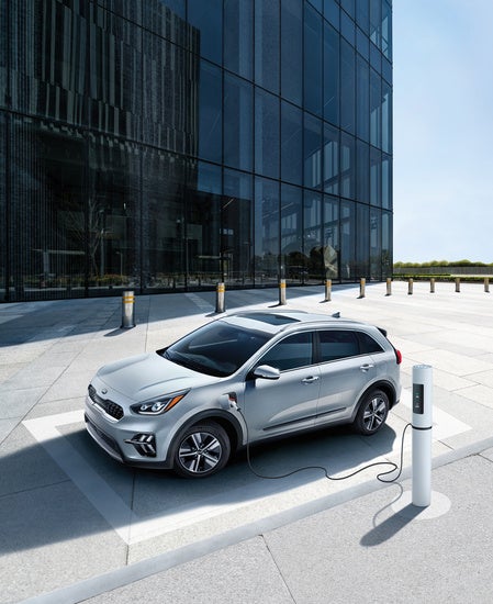 Kia K5 parked and charging at an electric vehicle station in front of a modern glass building on a sunny day.