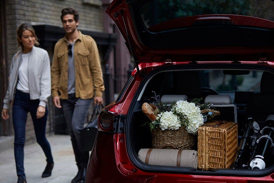 A man and a woman walk toward the open trunk of a red Mazda SUV which is packed with picnic items including wicker baskets and flowers.