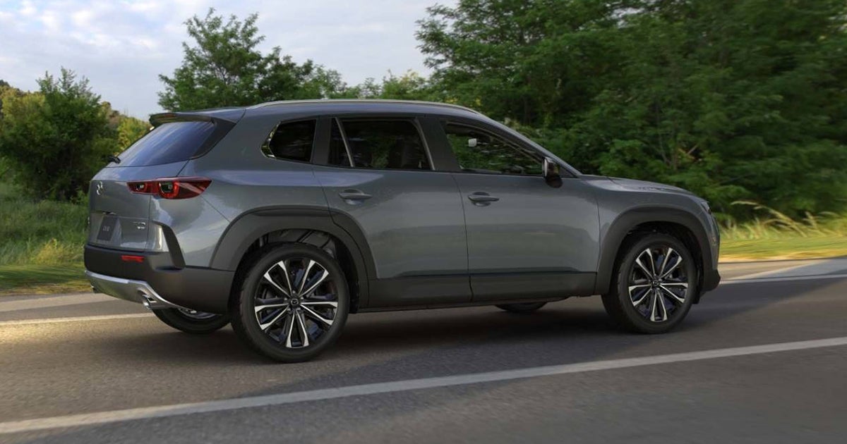 Gray Mazda CX-50 SUV side profile driving on tree-lined road