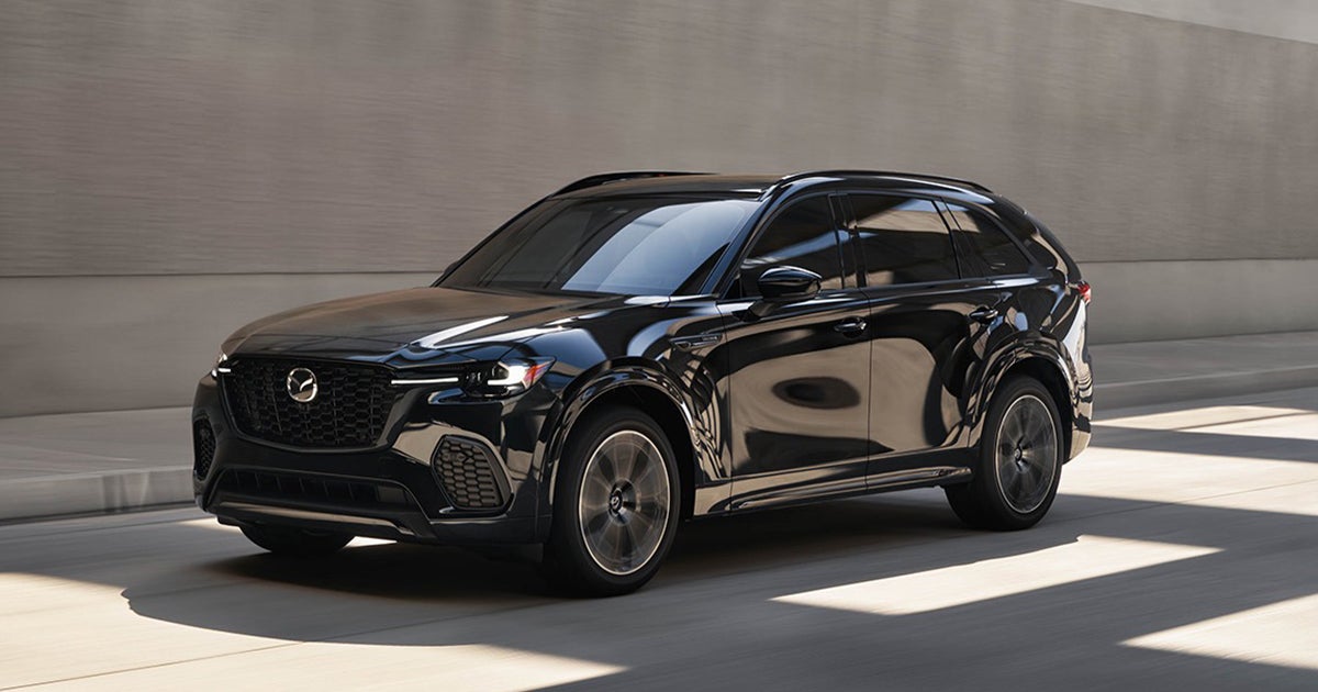 Black Mazda CX-90 SUV driving through a modern city street with sunlight reflecting off its glossy paint.