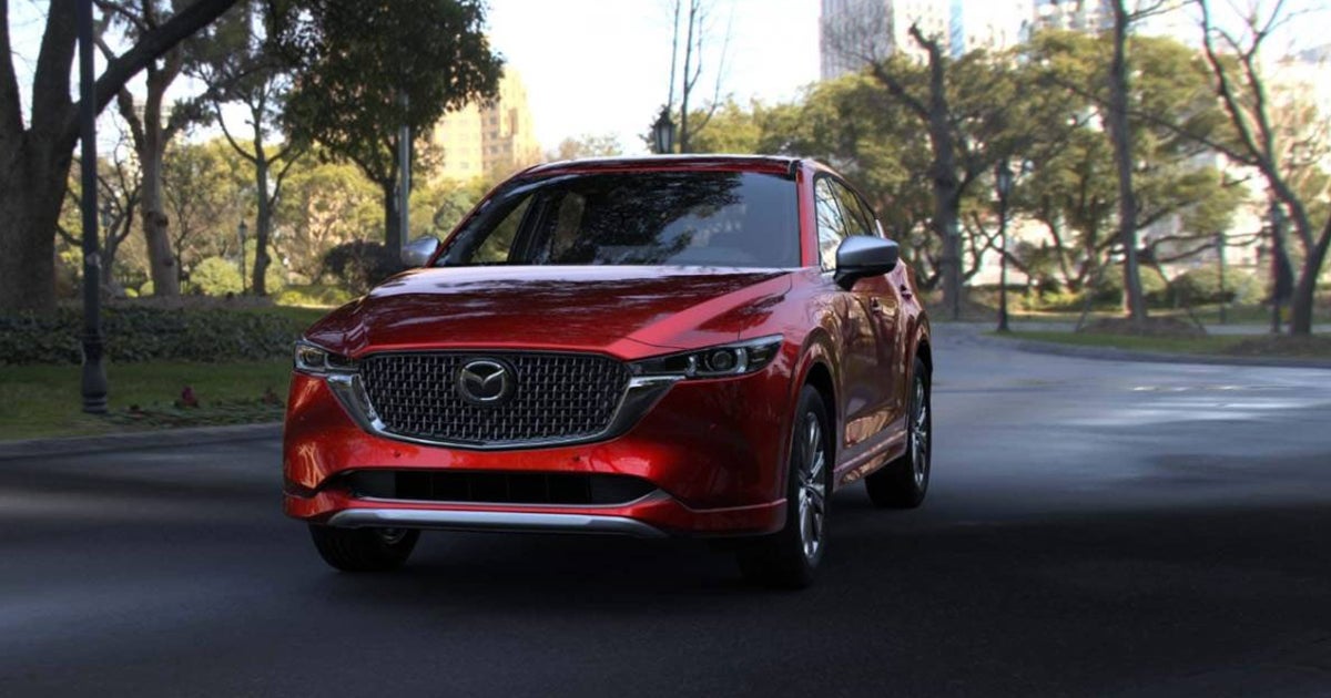 Red Mazda CX-5 SUV driving on a road lined with trees in an urban park setting.
