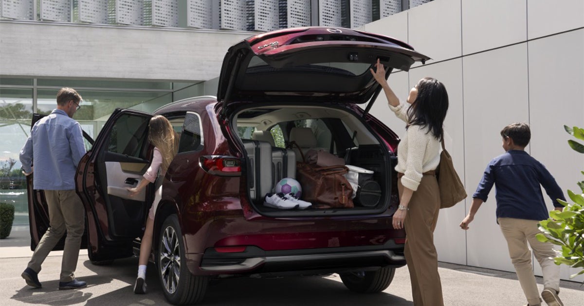 Mazda CX-90 SUV with a family loading luggage and gear into the open rear hatch outside a modern building.
