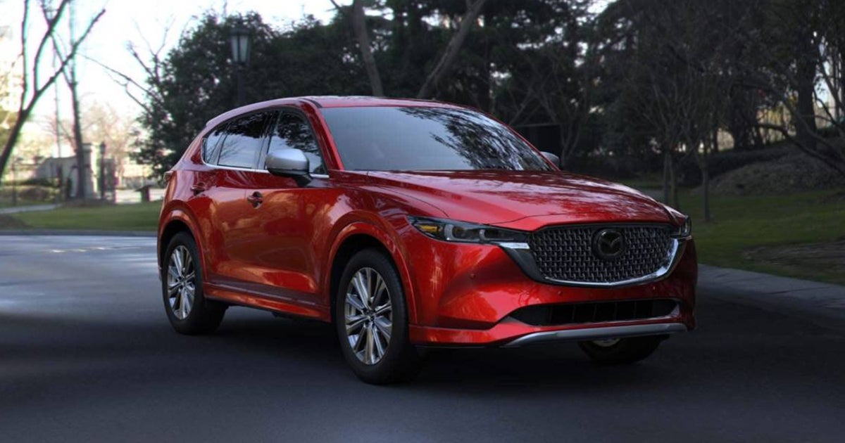 Red Mazda CX-5 SUV viewed from the front driver side while driving along a city street with trees in the background.