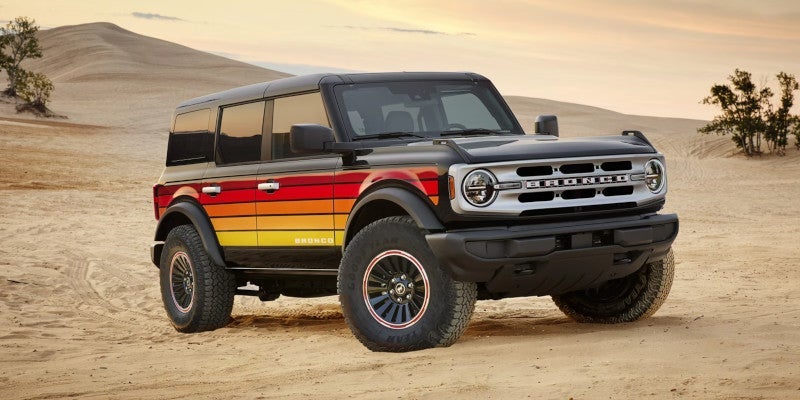 Black Ford Bronco with retro red, orange, and yellow stripes on its side, parked on sand dunes.