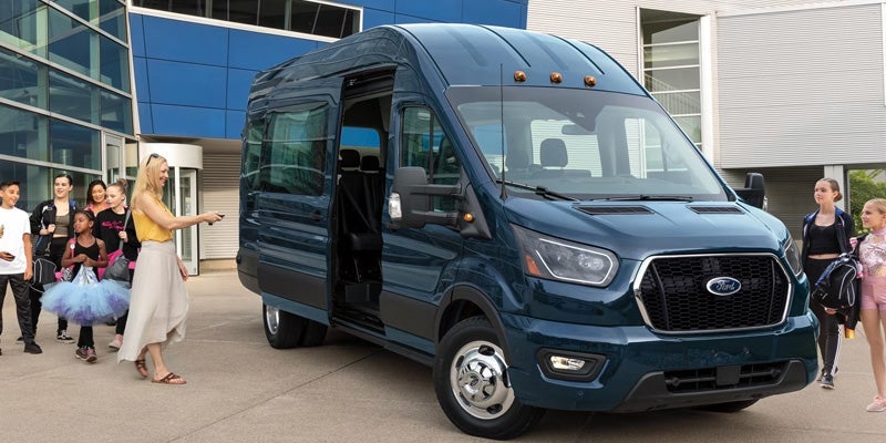 Dark blue Ford Transit passenger van with its sliding door open, a woman holding a remote, and a group of young dancers.