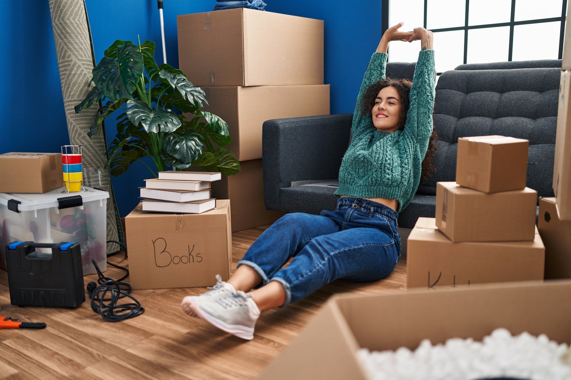 Young hispanic woman sitting on floor stretching arms at new home after moving.