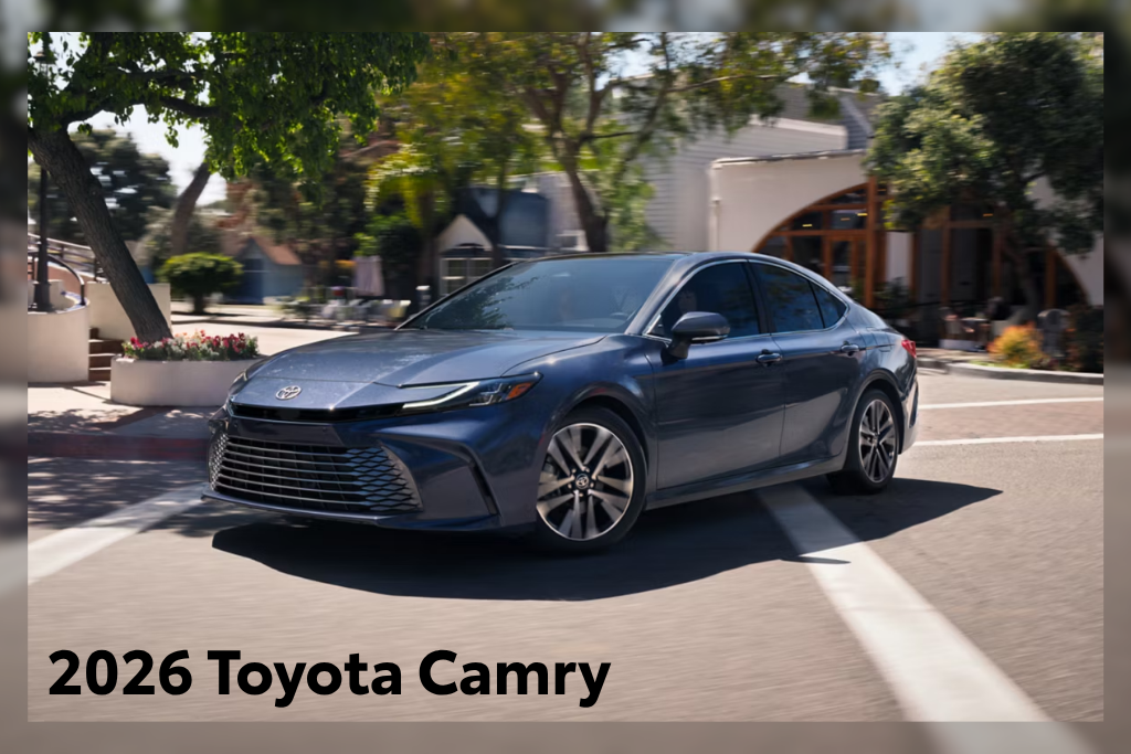 Blue 2026 Toyota Camry sedan driving through a sunny suburban street with trees and storefronts in the background.