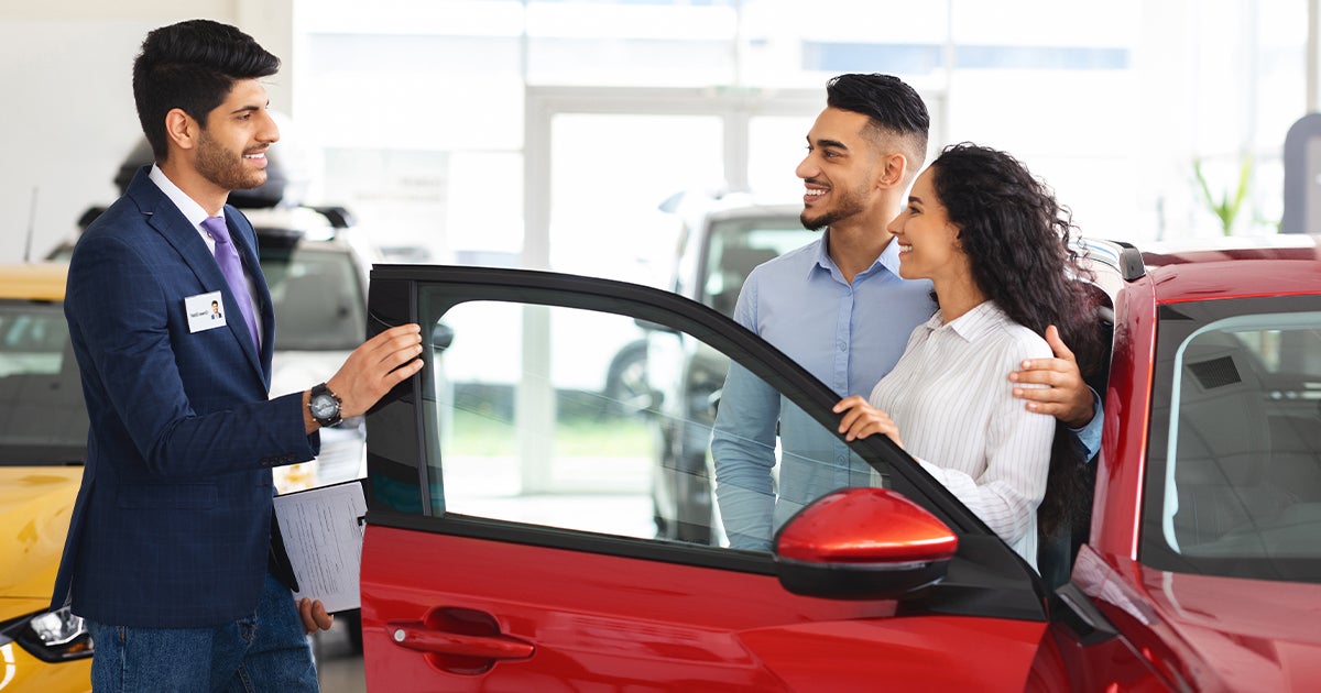 A couple leaning on a car in the car dealership while talking to a car salesperson.