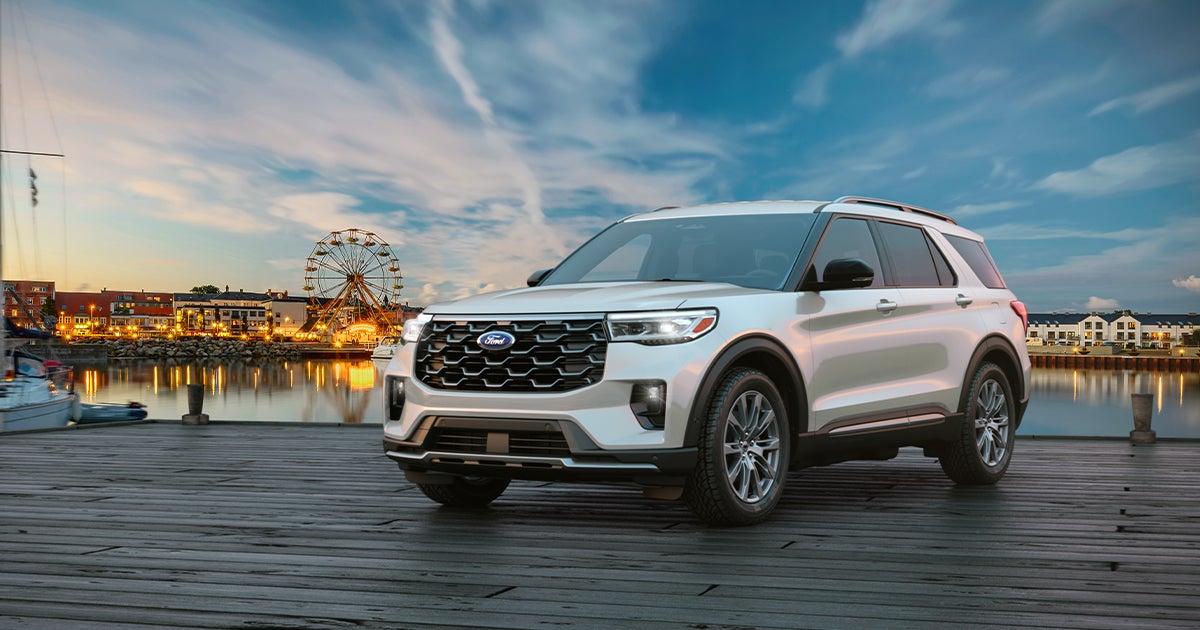 A white Ford Explorer midsize SUV parked on a wooden pier near a waterfront town.