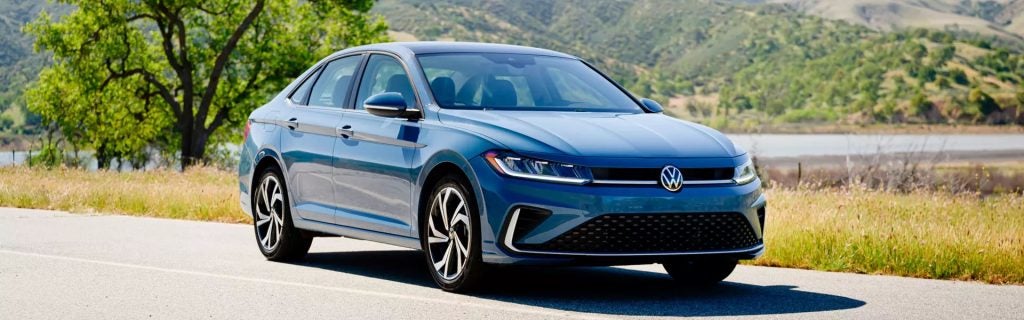 Blue 2026 Volkswagen Jetta driving on a scenic road with green hills in the background.