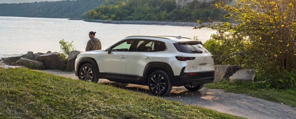 A Mazda CX-50 parked near a lake, with a person standing beside it, enjoying the scenic view.