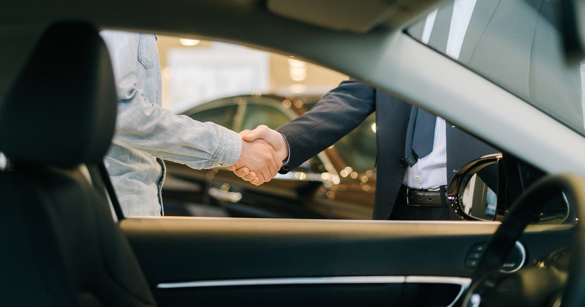 A car salesperson and customer shaking hands viewed from the inside of a vehicle at the car dealership.