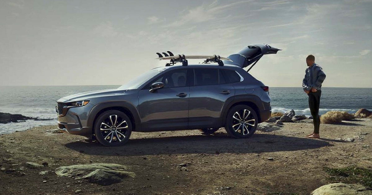 A Mazda CX-50 crossover SUV featuring a roof rack with surfboards parked by a beach.