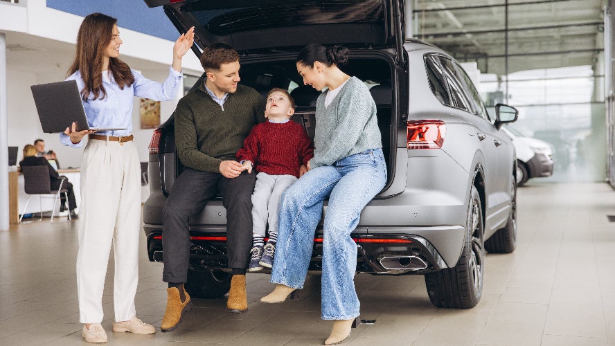 A family sitting in the trunk of an SUV at a dealership being shown it by a salesperson.