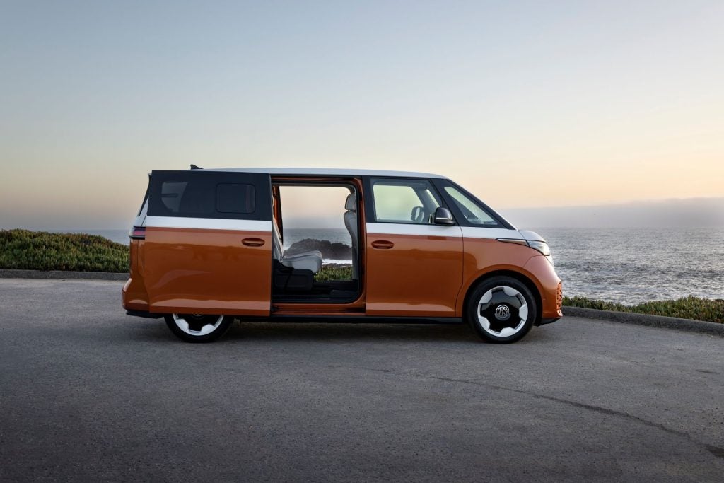 The Sleek Volkswagen ID. Buzz With Its Rear Doors Open, On Edge Of A Hill, With A Glittering Ocean In The Distance