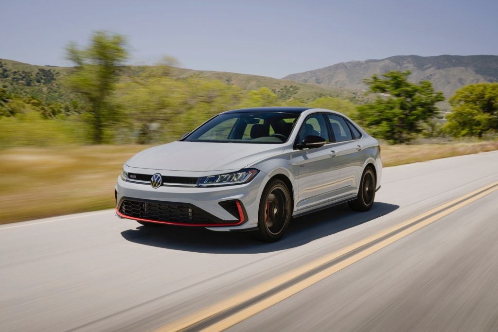 The Underrated And Affordable 2026 Volkswagen Jetta GLI Driving Down The Road, With Blurred Trees And Magnificent Mountains In The Background.
