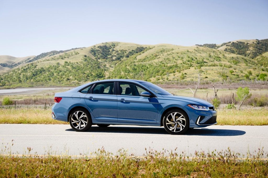 A Blue And Ventilated Volkswagen Jetta, Leisurely Driving Past Beautiful Summer Greenery
