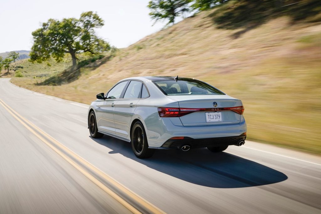 The Backside Look At The Magnificent 2026 Volkswagen Jetta GLI As The Car Drives Past High Hills And Trees Spread Out On The Plains