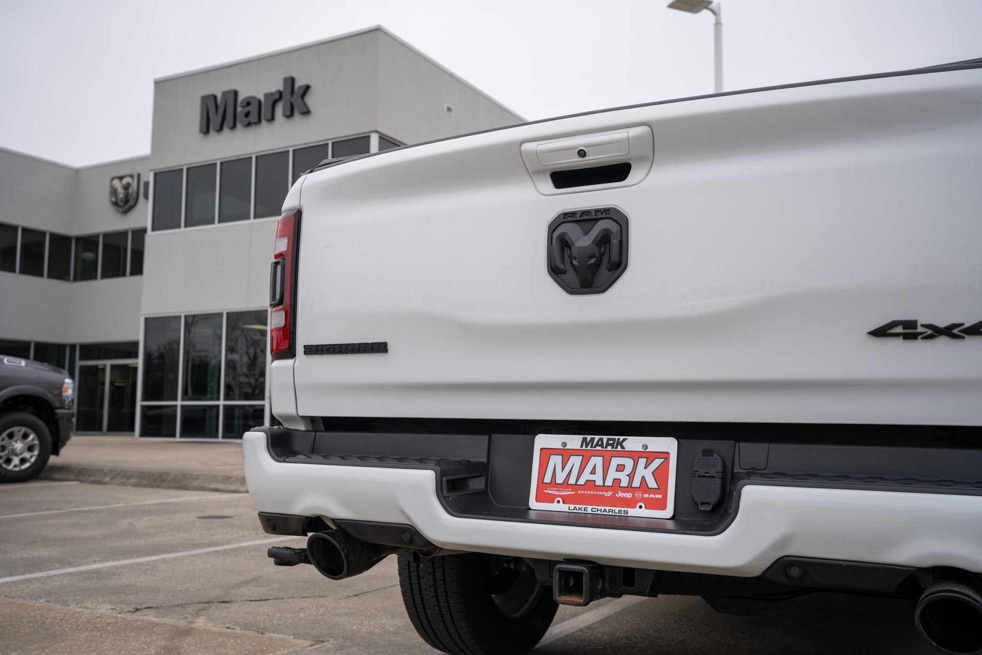 Close-up rear view of a white Ram Bighorn 4x4 pickup's tailgate and Mark Dodge dealer license plate, with the dealership building in the background.