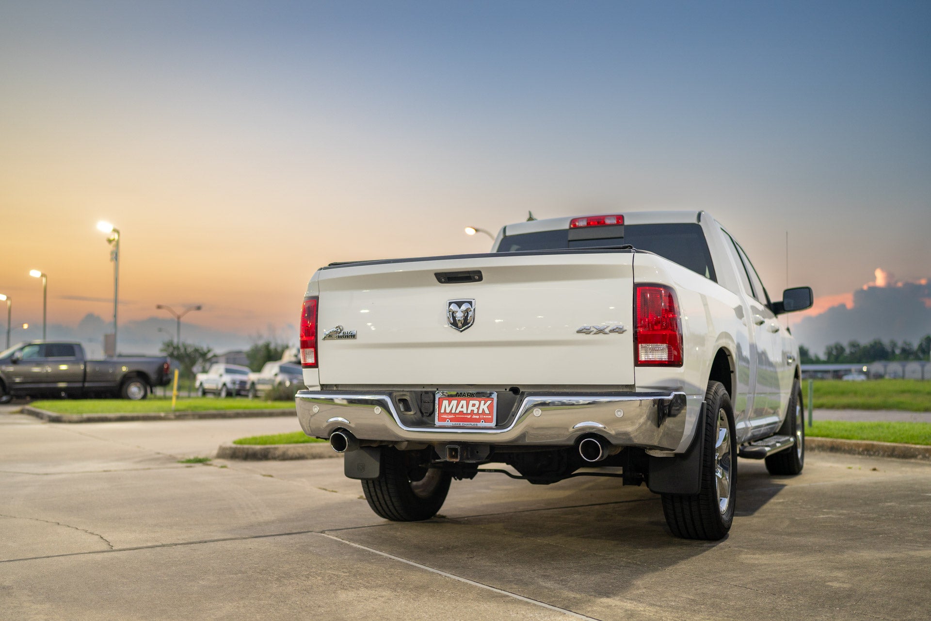 Rear view of a white Ram 1500 Laramie pickup with a Mark Dodge dealer plate, parked at sunset in the dealership lot with other trucks visible.