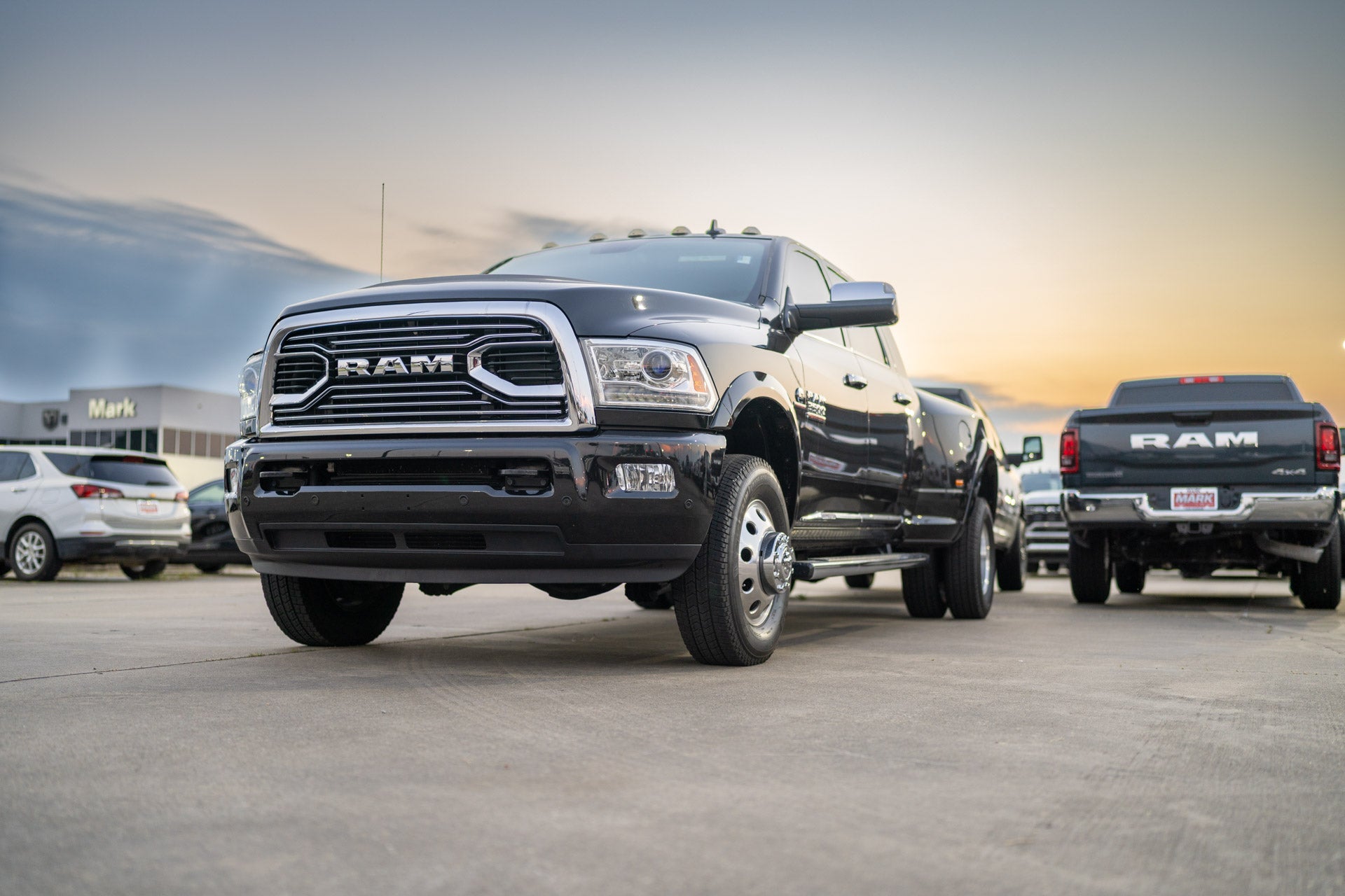 Front three-quarter view of a black Ram 3500 dually pickup at dusk, with another Ram truck and the Mark Dodge building in the background.
