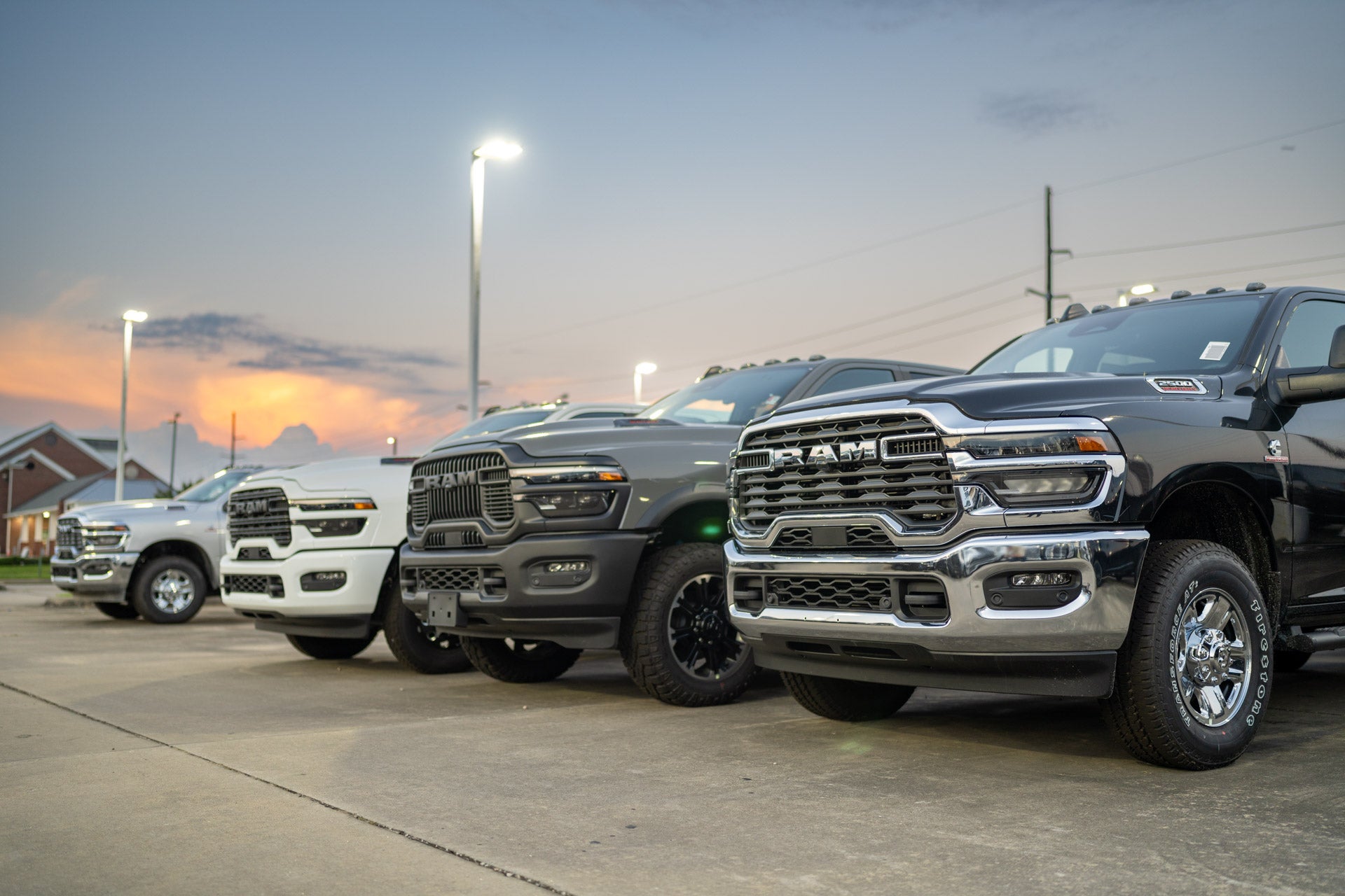 Row of Ram pickup trucks (white, dark gray, black) parked at sunset in the dealership lot, dramatic orange sky in background.