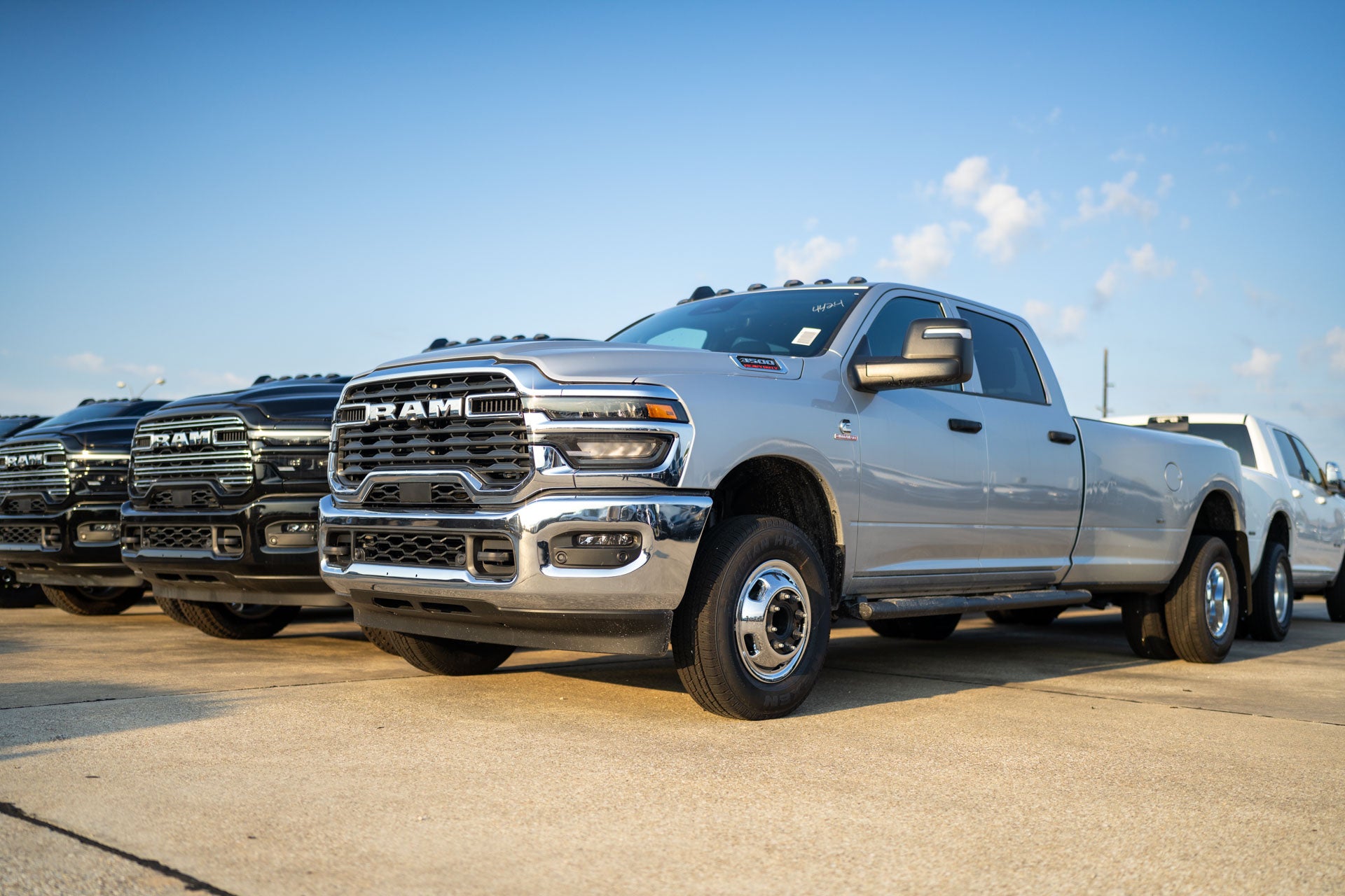 Close-up front three-quarter view of a silver Ram 3500 dually pickup, with a row of black Ram trucks and a white truck visible behind it.