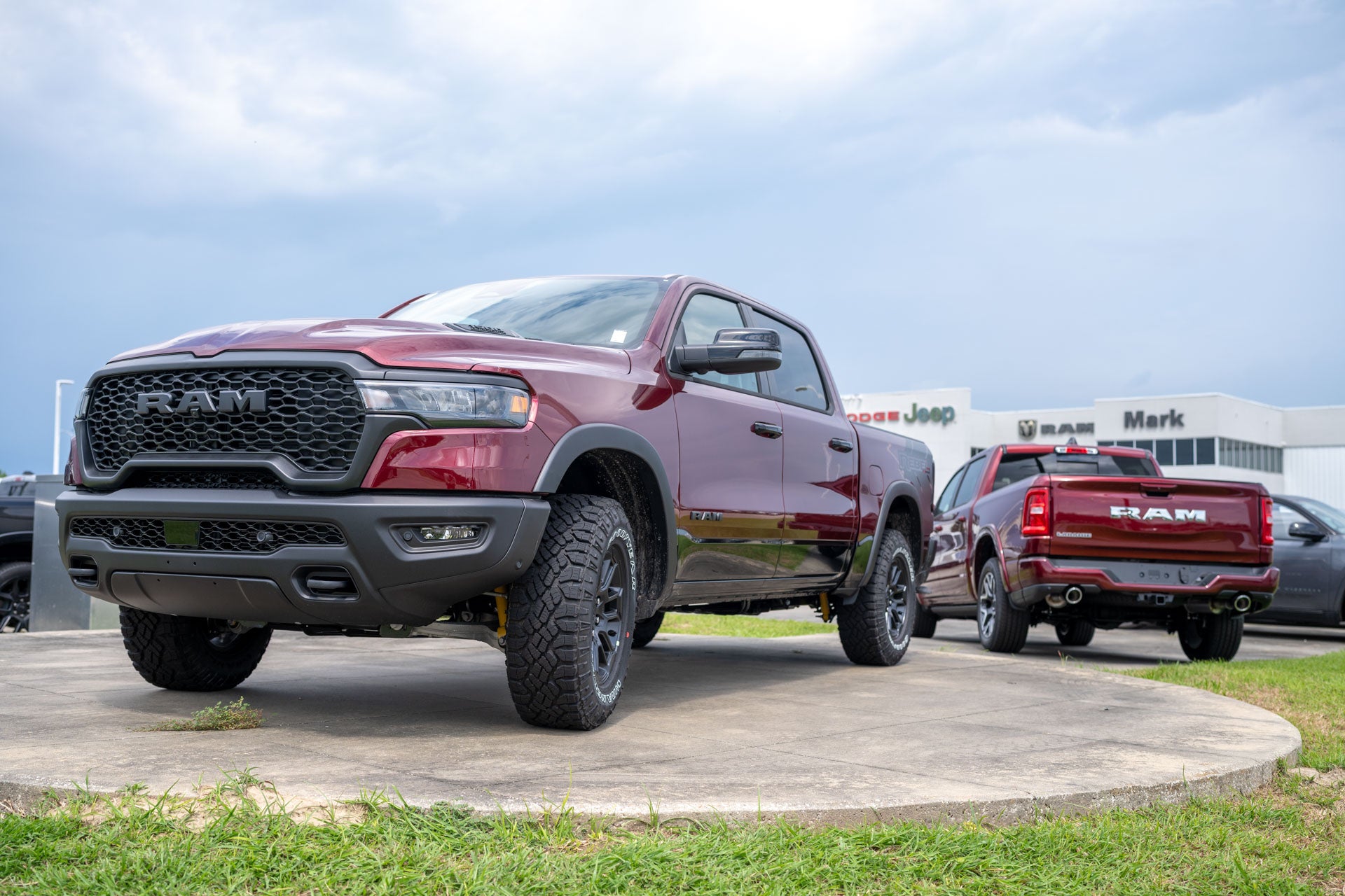 Front three-quarter view of a maroon Ram 1500 pickup on a display pedestal, with a second Ram truck and the Mark Dodge building behind.