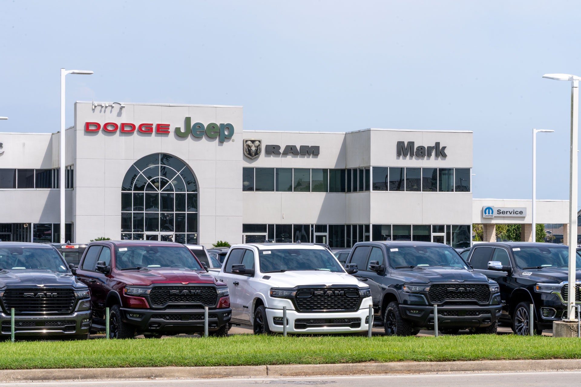 Row of Ram 1500 pickup trucks in maroon, white, gray, and black parked in front of the Mark Dodge Chrysler Jeep Ram dealership building.