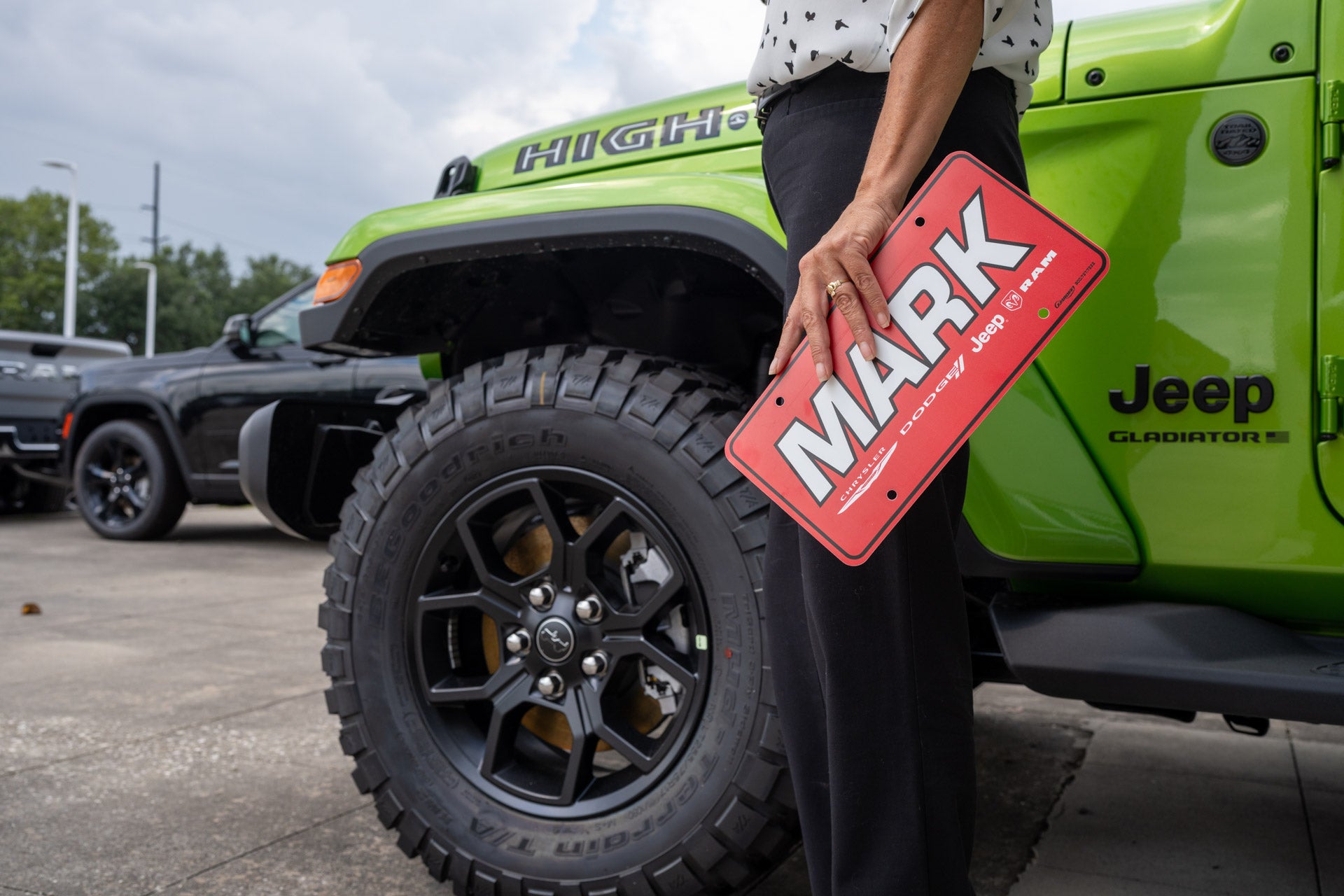 A hand holds a red MARK dealer license plate against the fender of a bright green Jeep Gladiator pickup.