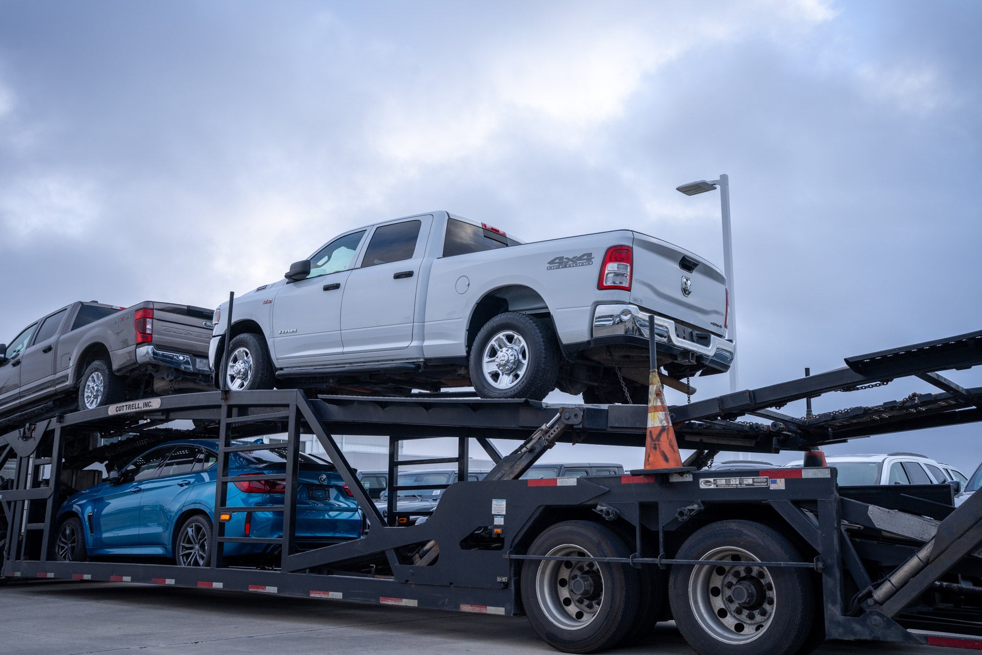A white Ram 1500 pickup loaded on a car-carrier delivery trailer, with a blue car on the lower deck.