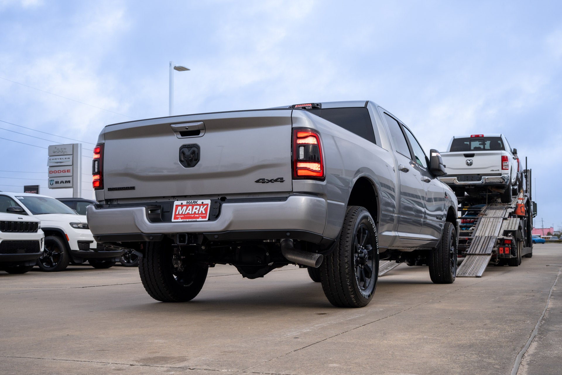 Rear three-quarter view of a silver Ram 2500 Bighorn pickup with a Mark Dodge dealer plate, dealership signage and a car-carrier trailer in the background.