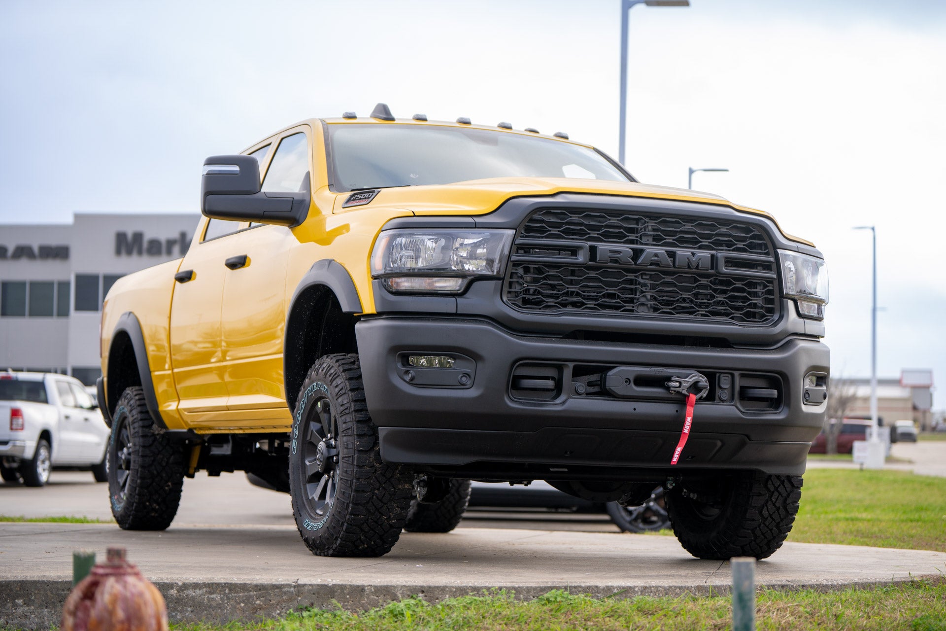 Front three-quarter view of a yellow Ram 2500 Power Wagon pickup with winch and roof-mounted lights, dealership building behind.