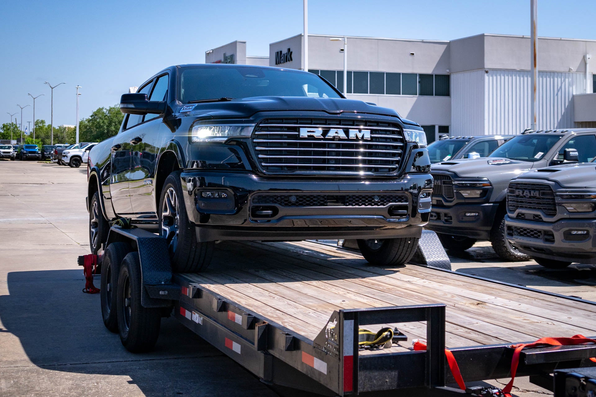 A black Ram 1500 pickup on a flatbed delivery trailer, with the Mark Dodge dealership building and Ram trucks visible in the background.