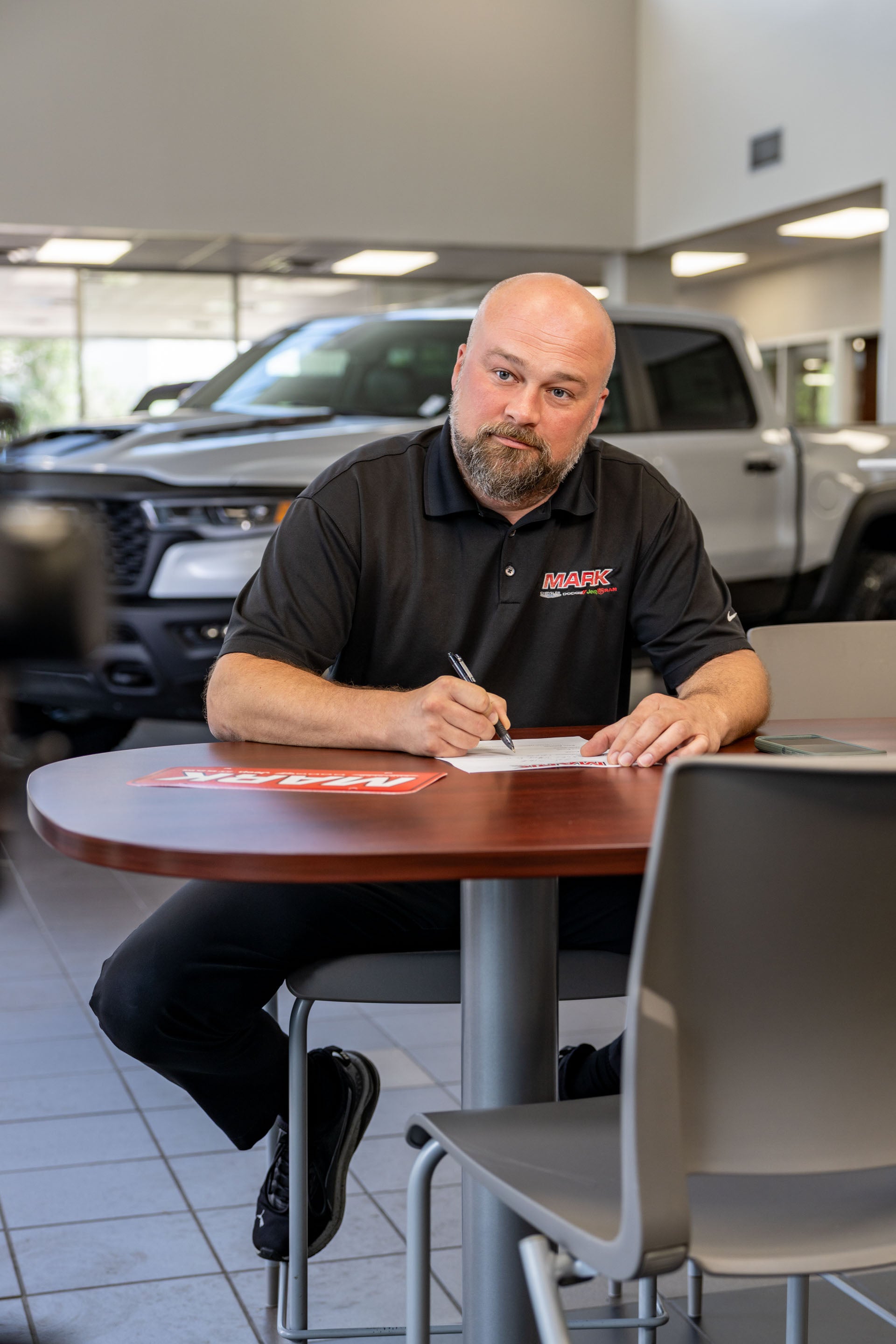 A Mark Dodge staff member in a black MARK polo sits at a table writing, with a white Ram TRX visible in the showroom behind him.