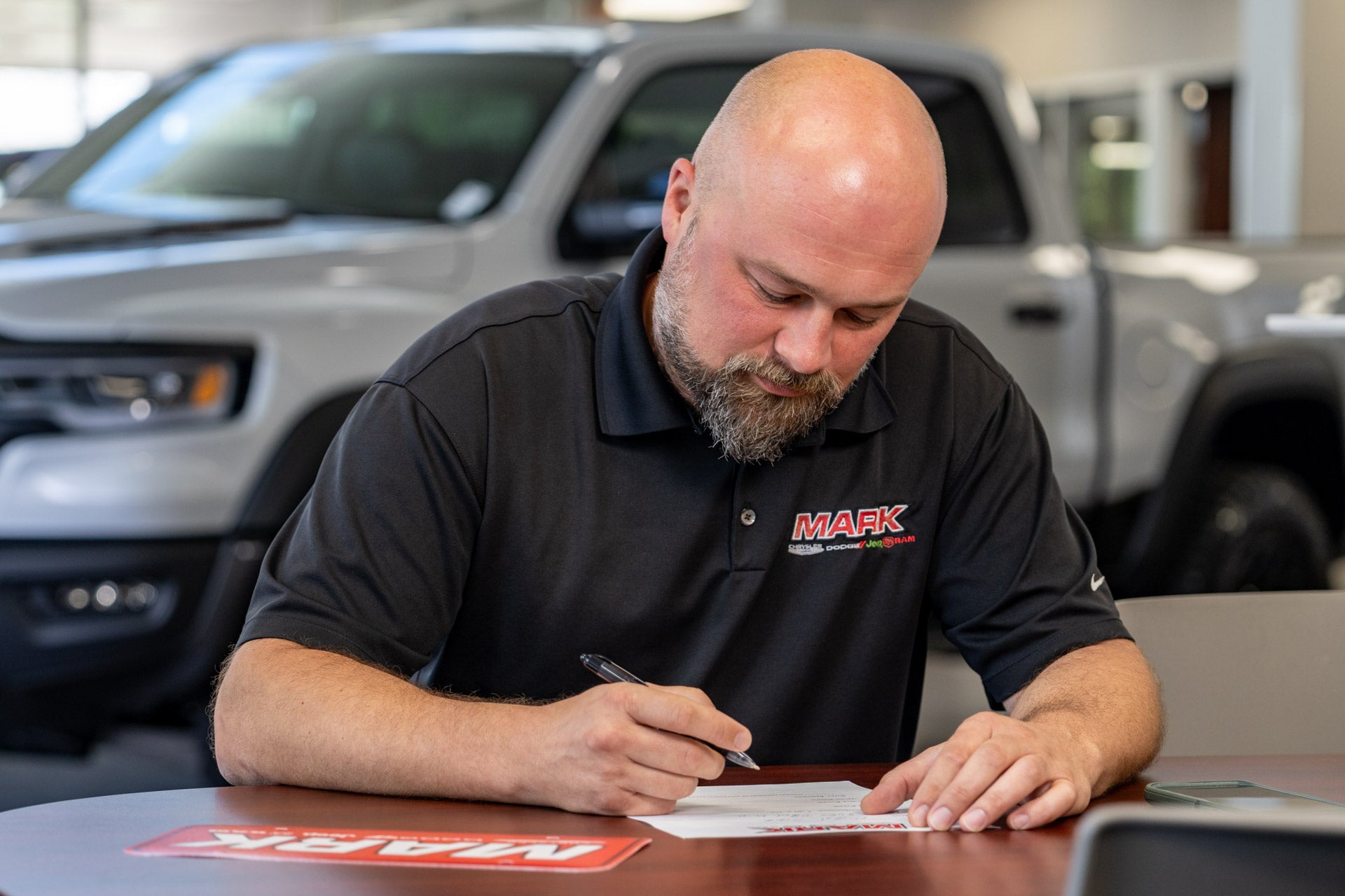 A Mark Dodge staff member in a black MARK polo writes on paper at a table, with a white Ram TRX visible in the showroom behind him.